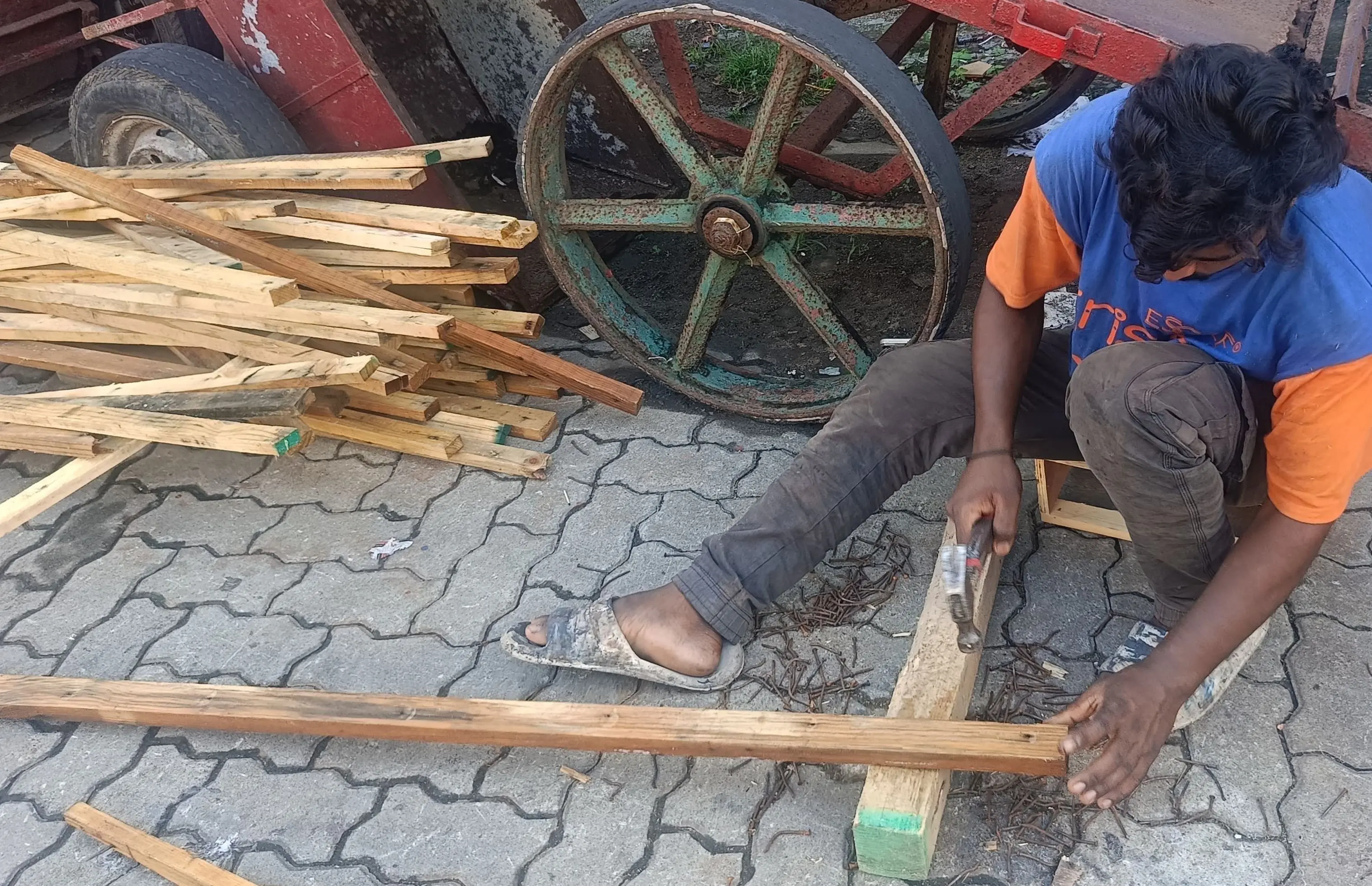 A person crouches on a paved surface, using a hand tool to work on a piece of wood resting on a small block. They wear a blue and orange shirt, dark trousers, and sandals. Behind them is a large pile of wooden planks and strips, and a vintage red cart with large metal-spoked wheels.