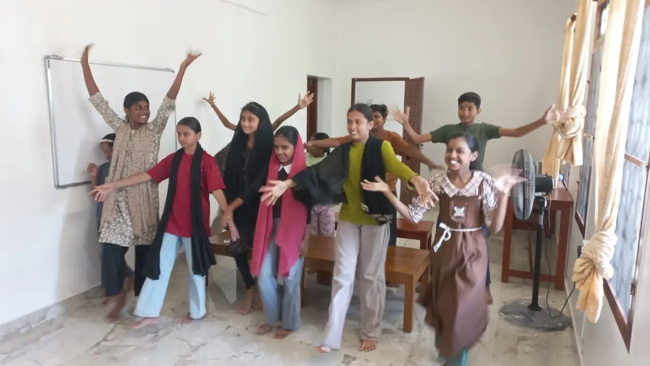 A group of children stands in a line inside a room, smiling and striking playful poses with their arms raised or extended. They wear a variety of casual clothing, including patterned tops, scarves, and dresses. Behind them, a whiteboard is mounted on the wall, and a doorway leads to another room. To the right, a standing fan sits on the floor near a table, and a window with tied-back curtains lets in light. The floor is tiled, and the walls are plain.