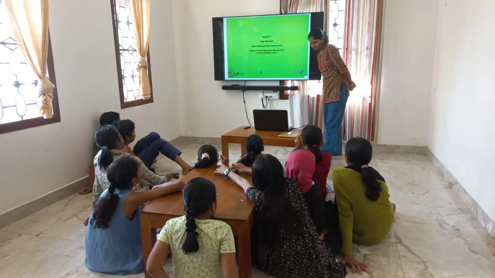 A group of children sit on the floor around a low wooden table in a room, facing a large screen mounted on the wall that displays a green background with text. An adult stands beside the screen, looking down at it. A laptop rests on the table beneath the screen. The room has windows with patterned glass and light curtains, and the floor is tiled.