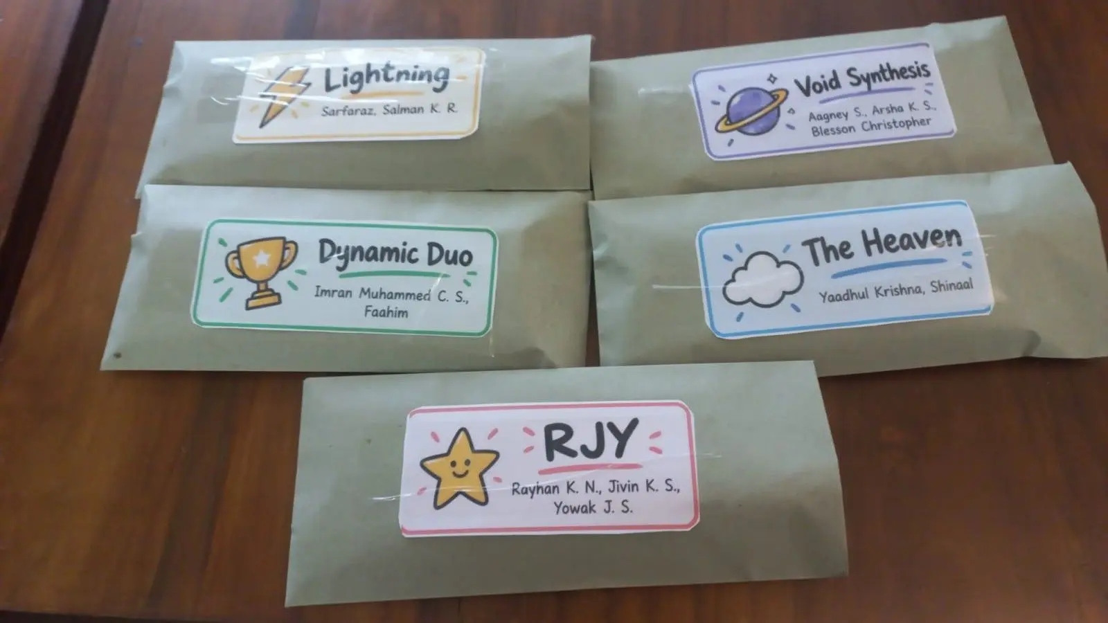 Five brown paper packages are arranged on a wooden surface, each sealed with clear tape and topped with a rectangular label. The labels feature different designs and names, including one with a lightning bolt, one with a planet, one with a trophy, one with a cloud, and one with a star. Each label includes a title and a list of names below it.