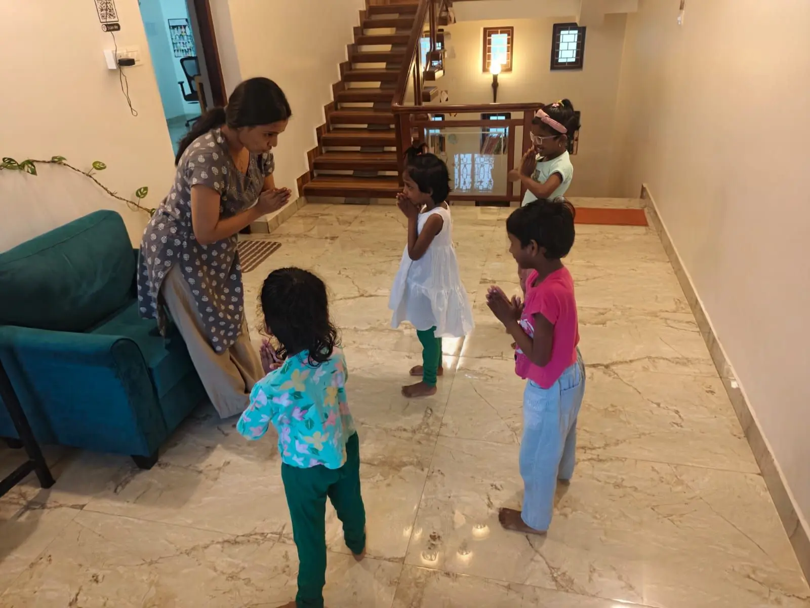 A woman stands beside a teal sofa, facing a group of five young children who are standing in a line on a tiled floor. All of them have their hands pressed together in front of them. Behind the children, a wooden staircase ascends against a wall, with a glass railing and framed pictures visible on the upper level.