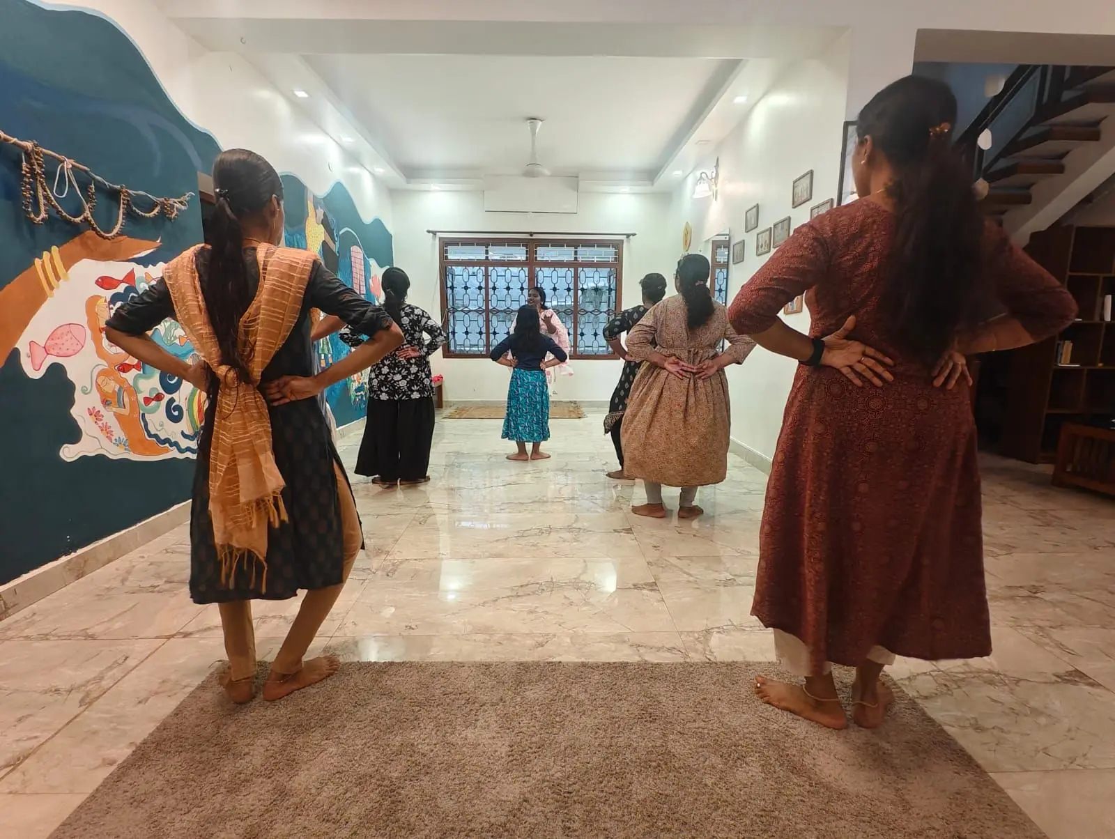 A group of people stand in a line inside a room with tiled floors and white walls, all facing away from the camera with their hands on their hips. On the left wall is a large painted mural with blue and orange colors and decorative elements hanging above it. At the far end of the room, a window with a grid pattern lets in light, and a person stands near it. To the right, framed pictures hang on the wall above a wooden bookshelf, and a staircase is visible in the background. A light-colored rug covers part of the floor in the foreground.