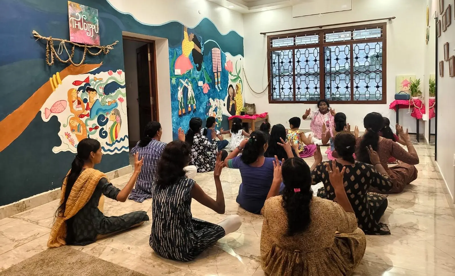 A group of people sit cross-legged on a tiled floor in a room with painted walls, facing a person who is seated at the front and gesturing with raised hands. The participants, mostly women and children, mirror the gesture with their own hands raised. The walls feature colorful murals, including a large painted hand and figures, and a doorway is visible on the left. A window with a decorative grille is on the back wall, and a small table with items on it stands to the right.