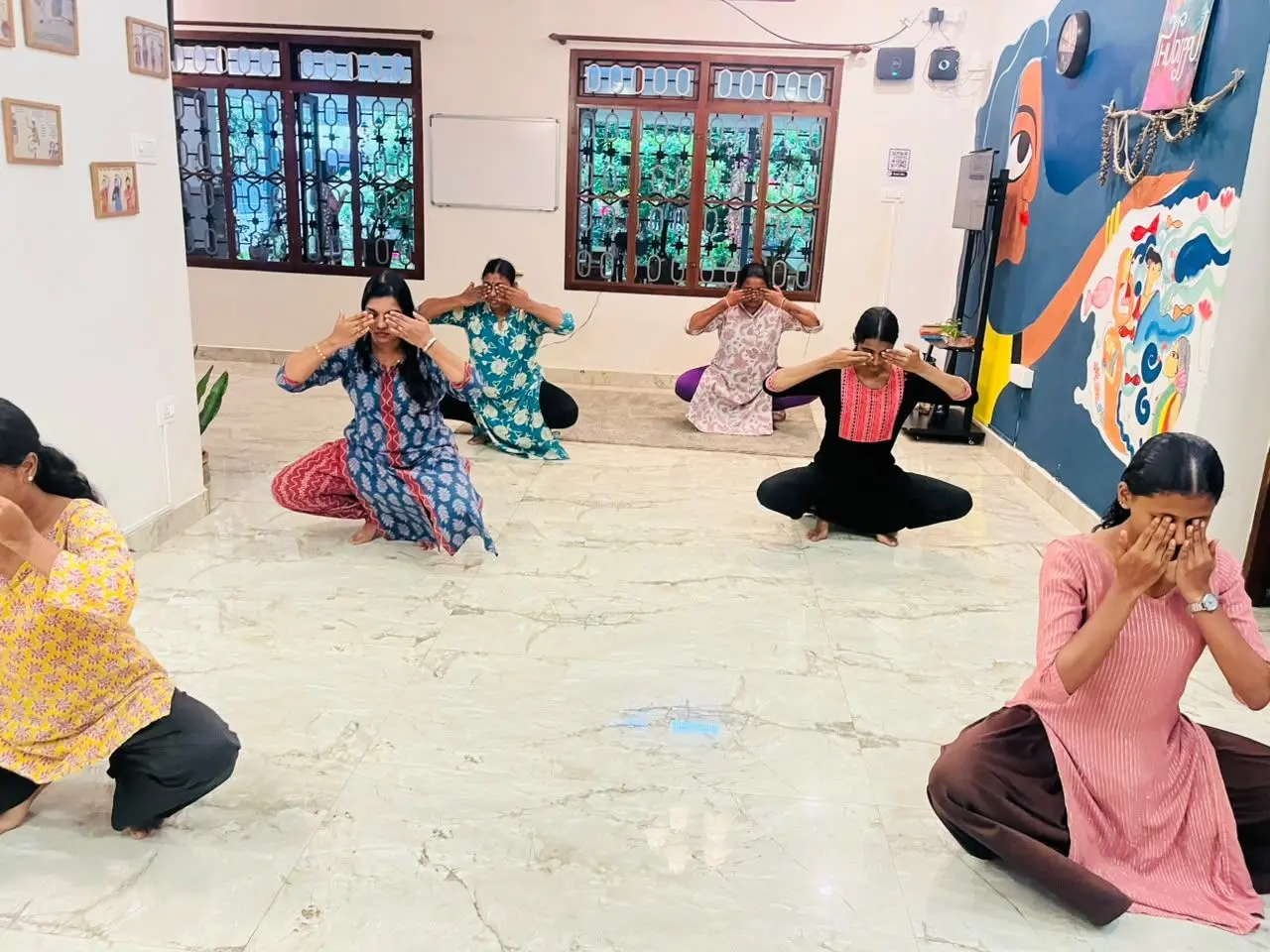 Six women are sitting on a tiled floor in a room, each with their hands covering their eyes. They are arranged in a loose semi-circle facing forward. The room has two large windows with decorative grilles, a whiteboard on the wall, and a colorful mural on the right side. The women are wearing various traditional outfits in different colors and patterns.
