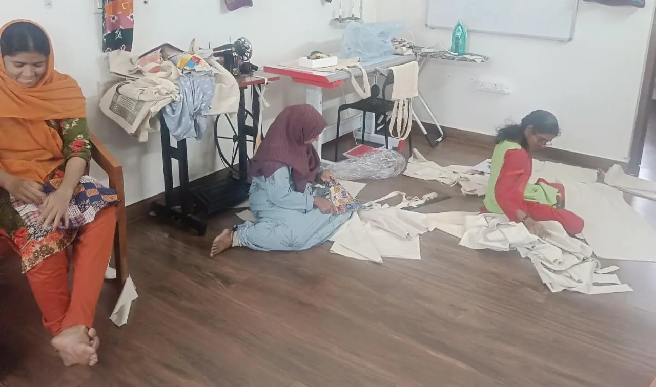 Three people are in a room with wooden flooring, surrounded by fabric pieces and sewing equipment. On the left, a person wearing an orange headscarf and matching outfit sits on a wooden chair, barefoot, with hands resting on their lap. In the center, another person in a maroon headscarf and light blue clothing sits on the floor, working with fabric. On the right, a third person in a green top and red pants sits cross-legged on the floor, also handling fabric. Behind them, a sewing machine sits on a stand with bags and materials nearby, and a table holds more supplies against a white wall.