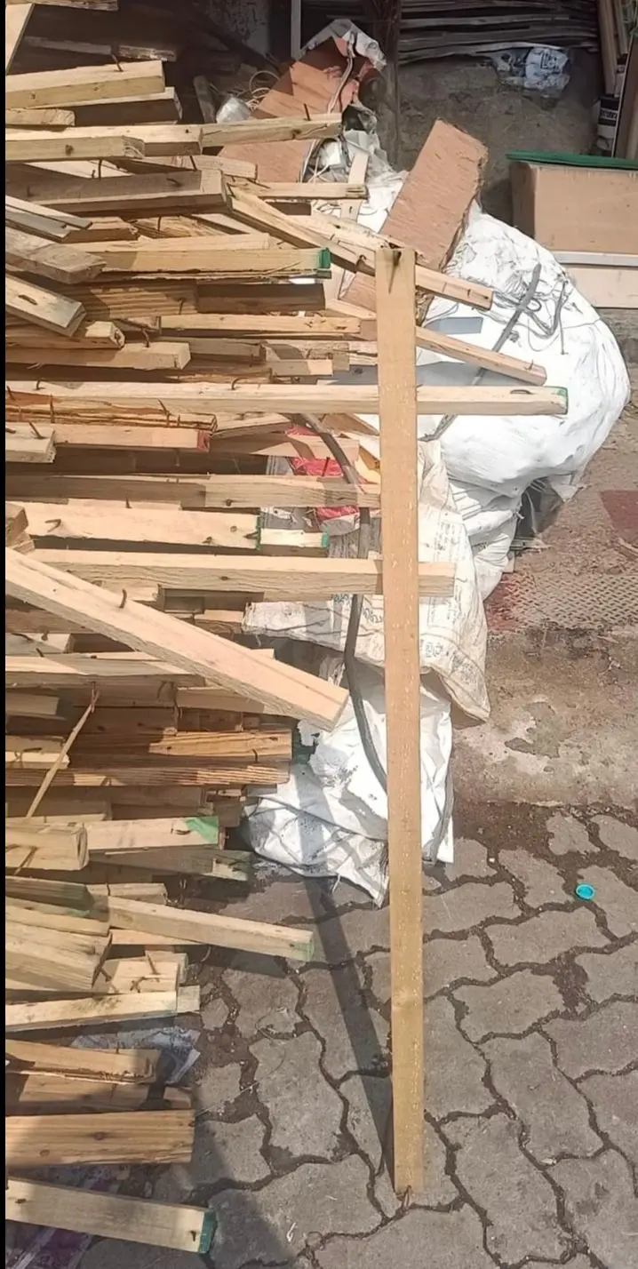 A large pile of scrap wooden planks and boards is stacked haphazardly on a paved surface, with a single long wooden plank standing upright in front of the pile. Behind the wood, white sacks and cardboard boxes are visible, along with other discarded materials. The ground is made of interlocking paving stones, and a small blue object lies on the pavement near the bottom right.