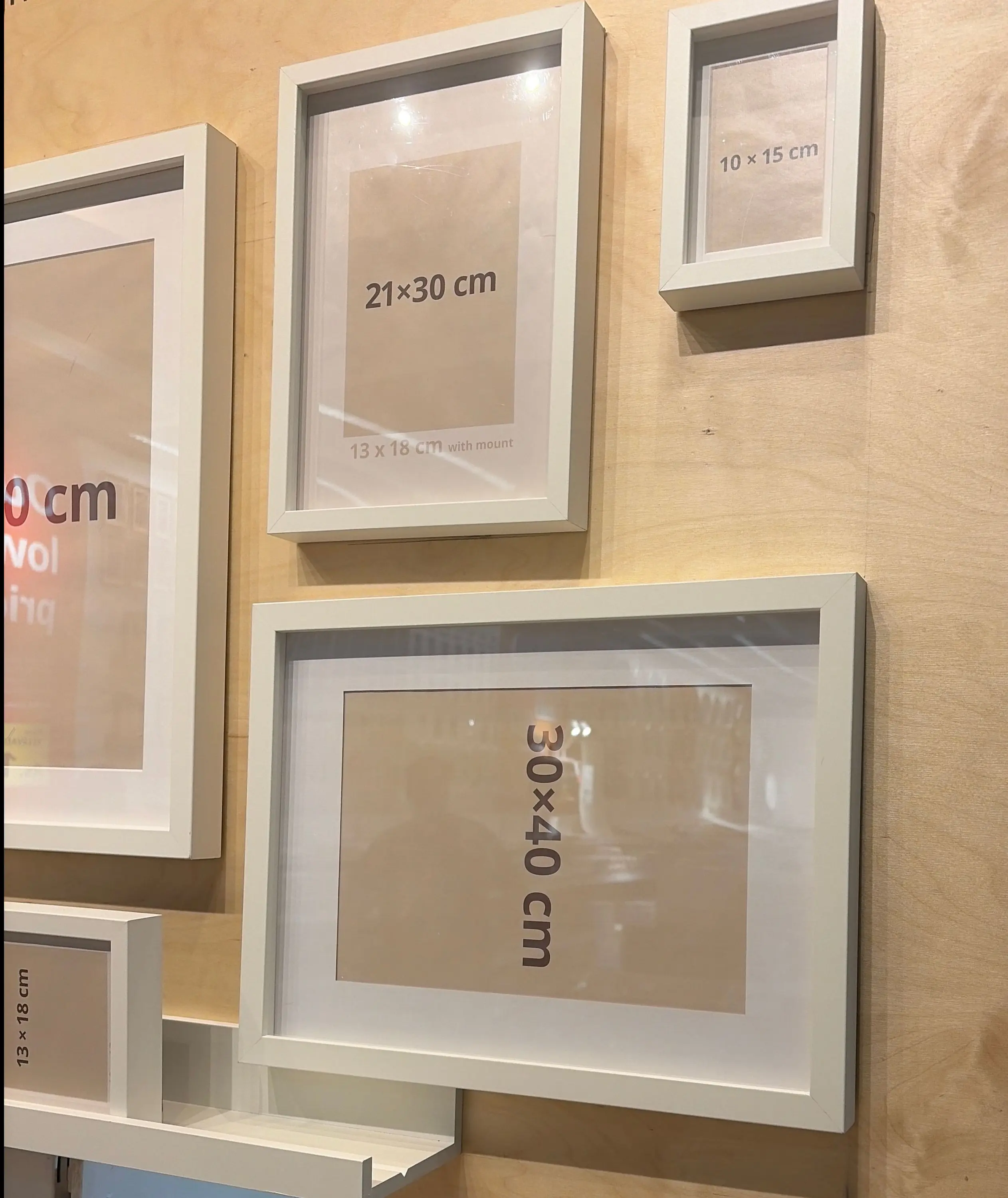 Several white picture frames of different sizes are mounted on a light wood wall, each containing a mat with printed dimensions indicating the frame size and compatible photo size.